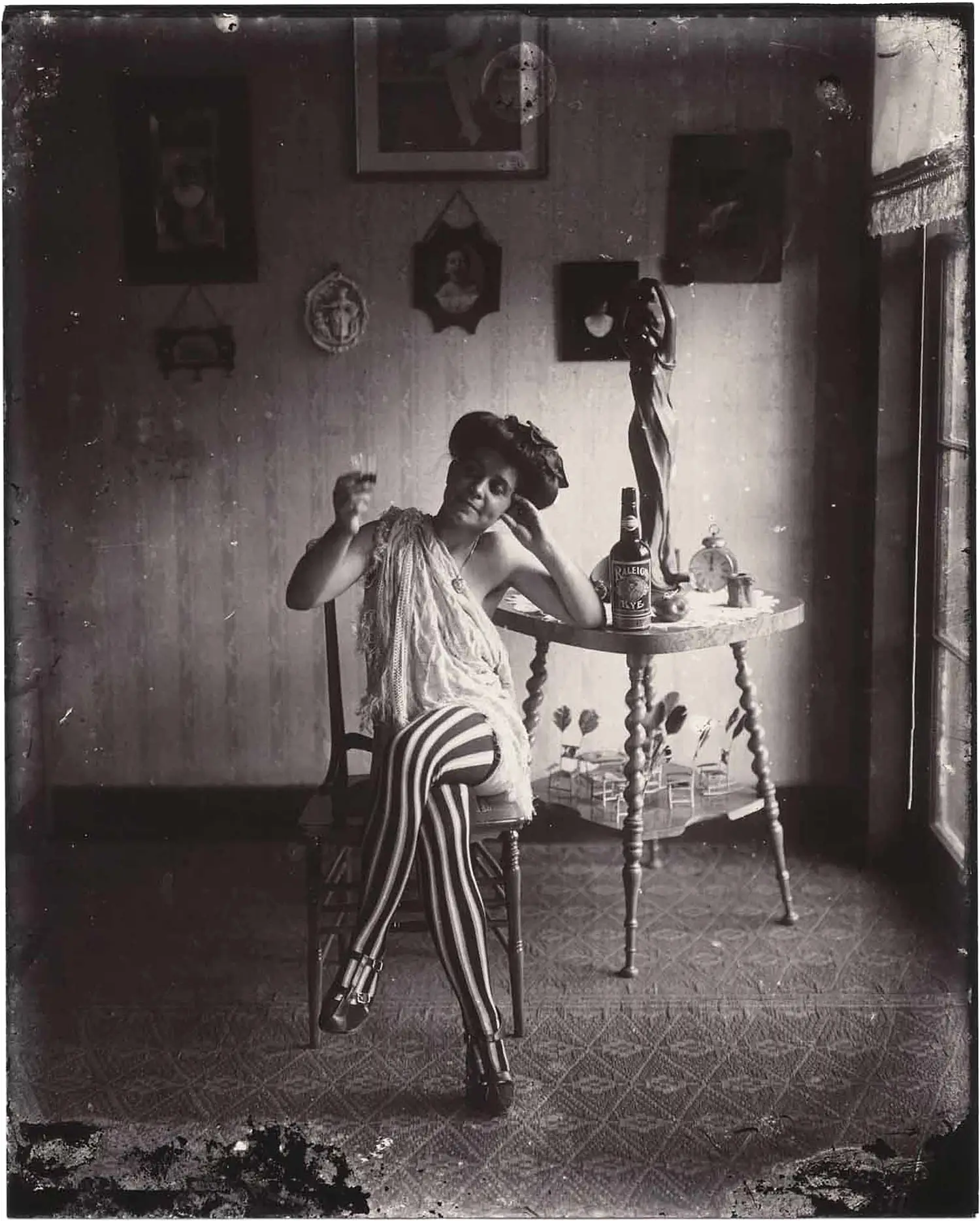 In this vintage photograph a woman is seated on a carved wooden chair. Her legs are crossed and she leans on a table next to her, upon which a bottle of rye is prominently placed. She is holding a partially filled glass with her right arm and is wearing black and white striped leggings and a dress that reveals her arms and left shoulder.